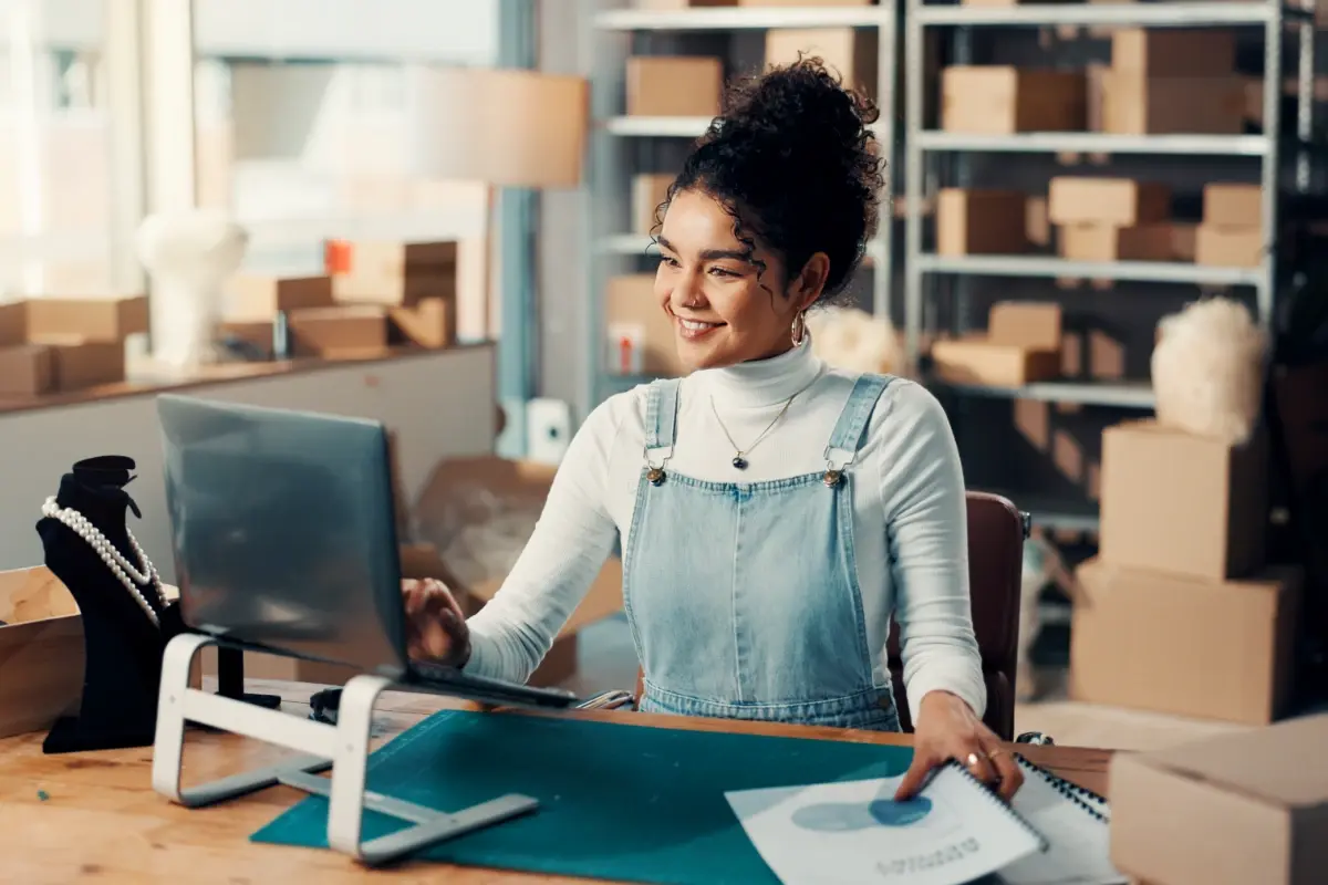 A smiling small business owner looks at her laptop screen while managing order fulfillment in her warehouse.