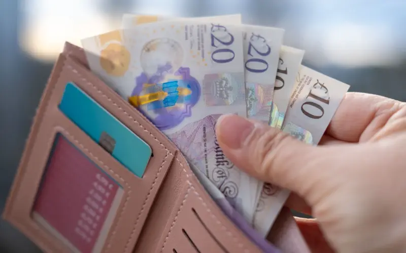 A hand pulling British pound sterling banknotes from a pink leather wallet.