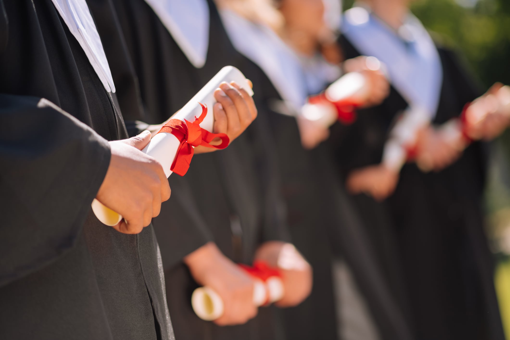 Close-up of graduates in black gowns holding rolled diplomas with red ribbons during a graduation ceremony.